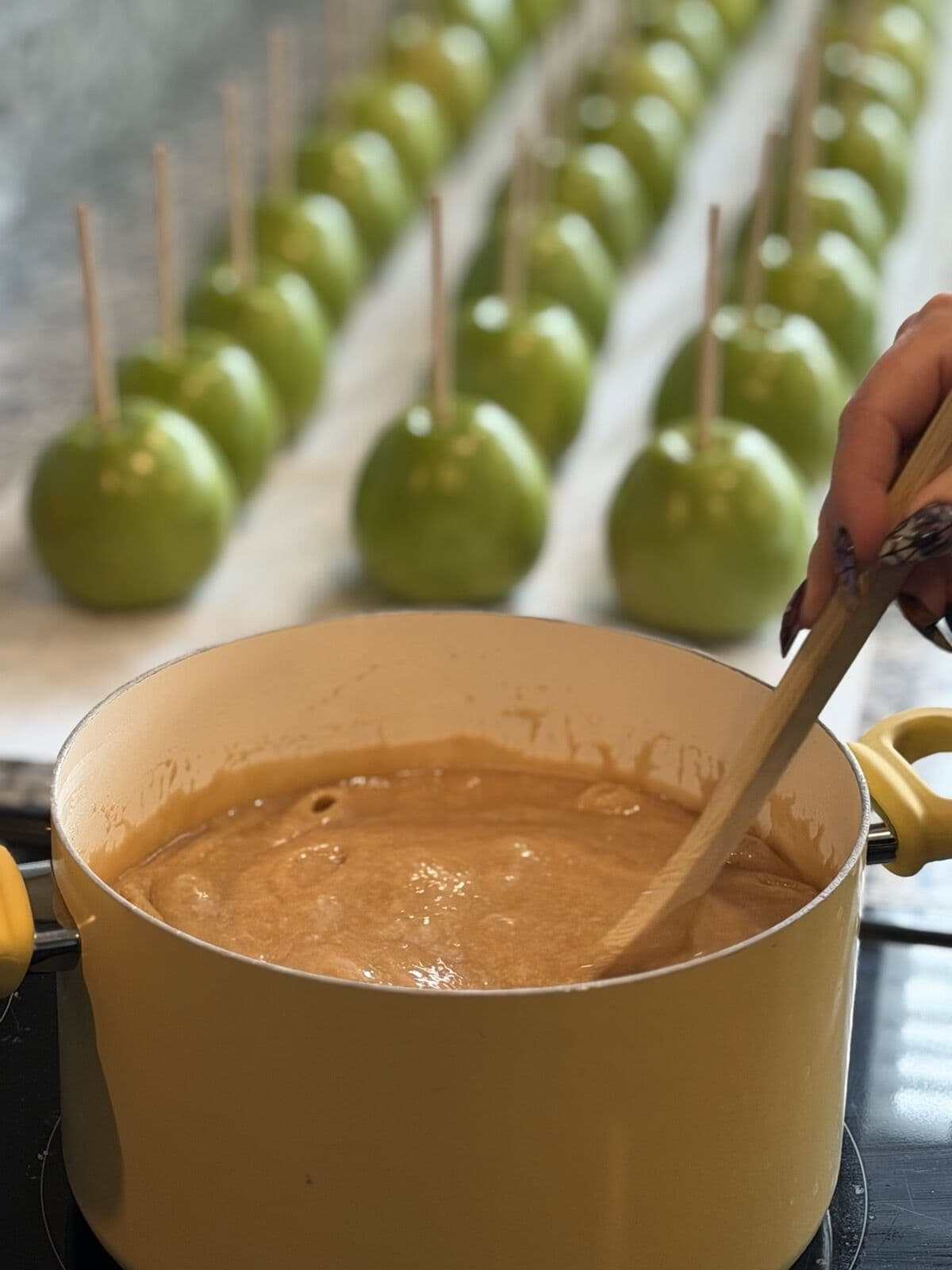 Stirring a pot of caramel with rows of green apples waiting to be dipped